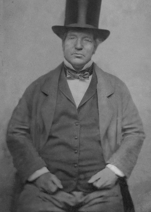 Black and white portrait of a man wearing a tall top hat, bow tie, and suit, seated with his hands resting on his thighs, facing the camera.