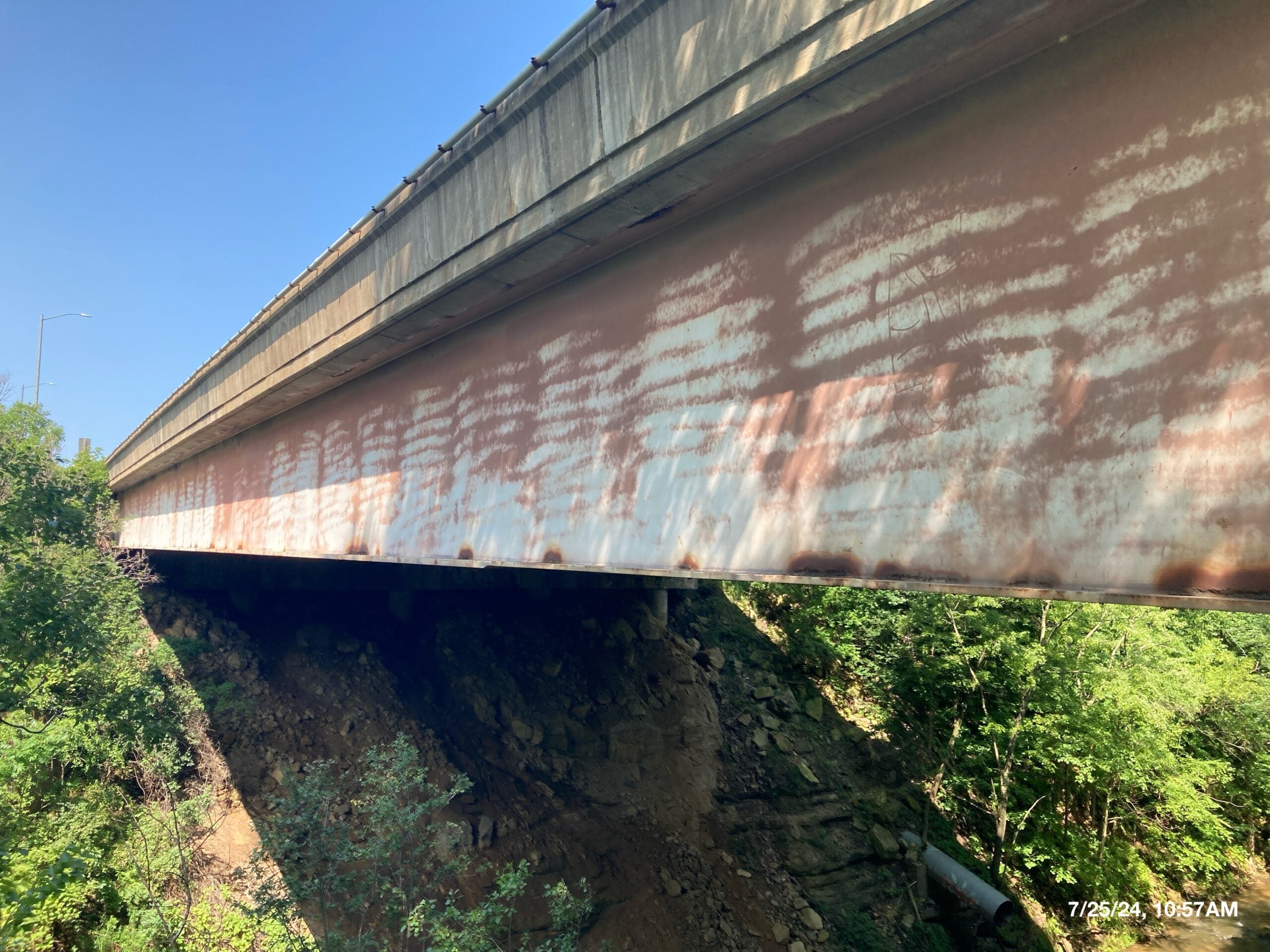A concrete and steel bridge with rust stains on its side spans over a rocky embankment with green vegetation below.