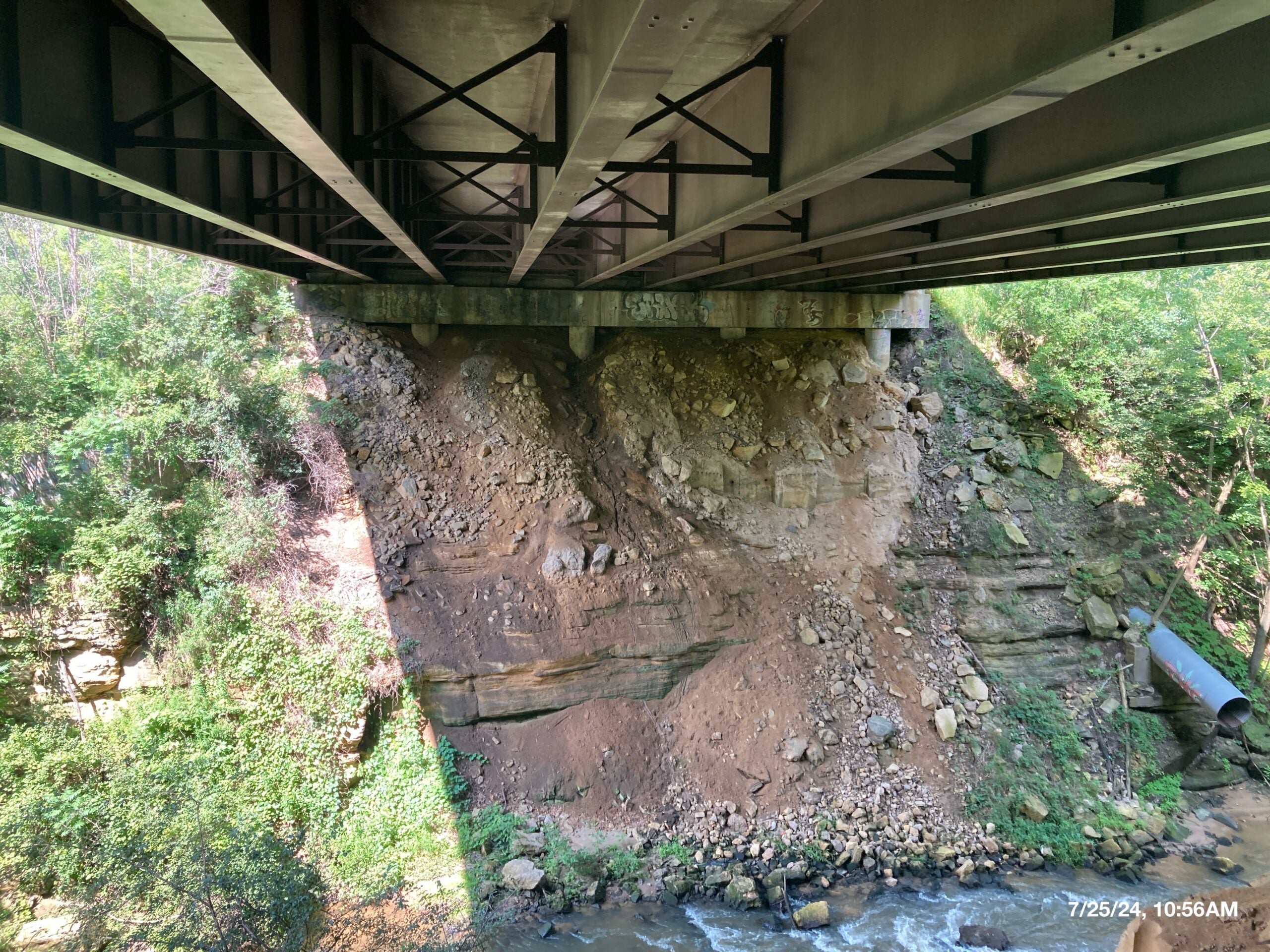 View from beneath a bridge showing an eroded, rocky embankment with exposed soil and scattered vegetation, overlooking a shallow stream below.