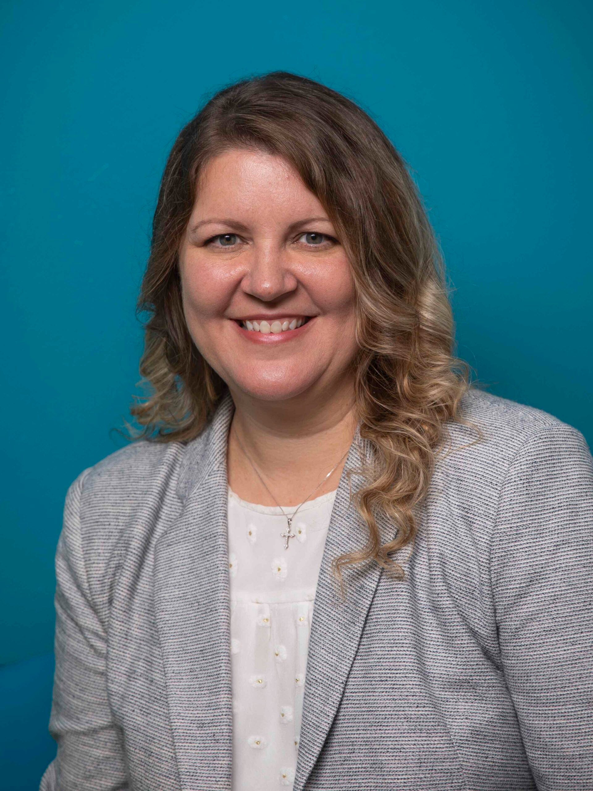 A woman with wavy hair wearing a light gray blazer and white blouse smiles in front of a solid blue background.