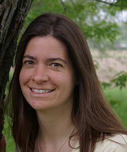 A woman with long brown hair and a light yellow shirt smiles outdoors near a tree, with green foliage in the background.