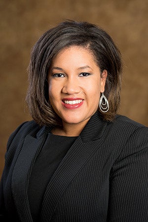 A woman with shoulder-length dark hair, wearing a black pinstripe blazer, black top, and silver earrings, smiles at the camera against a brown background.