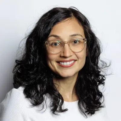 A woman with wavy dark hair, wearing clear-framed glasses and a white top, is smiling in front of a plain white background.