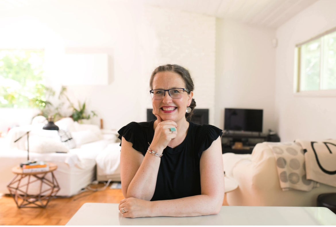 A woman wearing glasses and a black shirt sits at a table, smiling with her hand resting on her chin in a bright, modern living room.