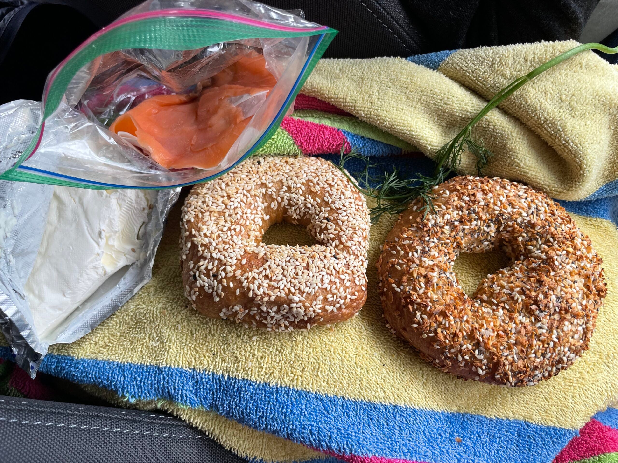 Two bagels, a container of cream cheese, and a bag of smoked salmon are arranged on a striped towel. Fresh dill is placed next to the bagels.