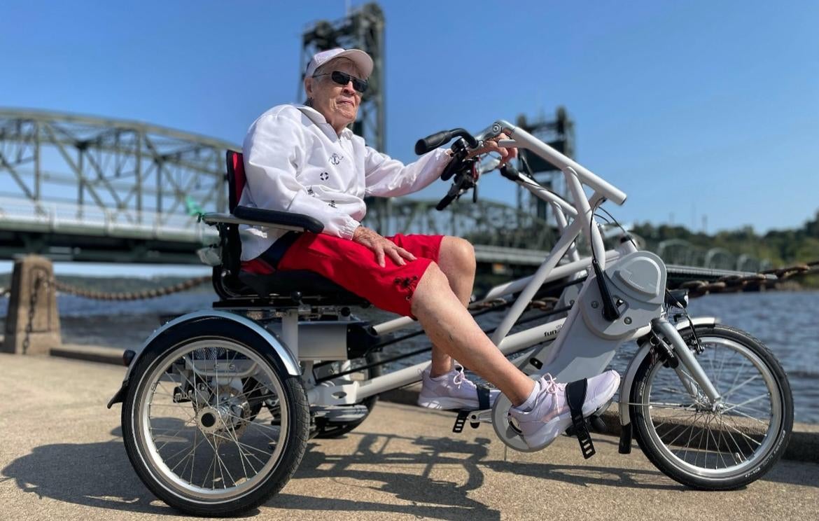 An older adult wearing a white cap and red shorts sits on a three-wheeled adaptive bicycle by a river, with a bridge in the background on a sunny day.