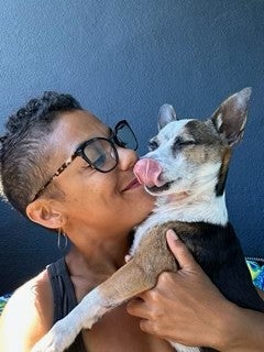 A person wearing glasses holds a small dog that is licking its nose, both appearing close and content against a dark background.