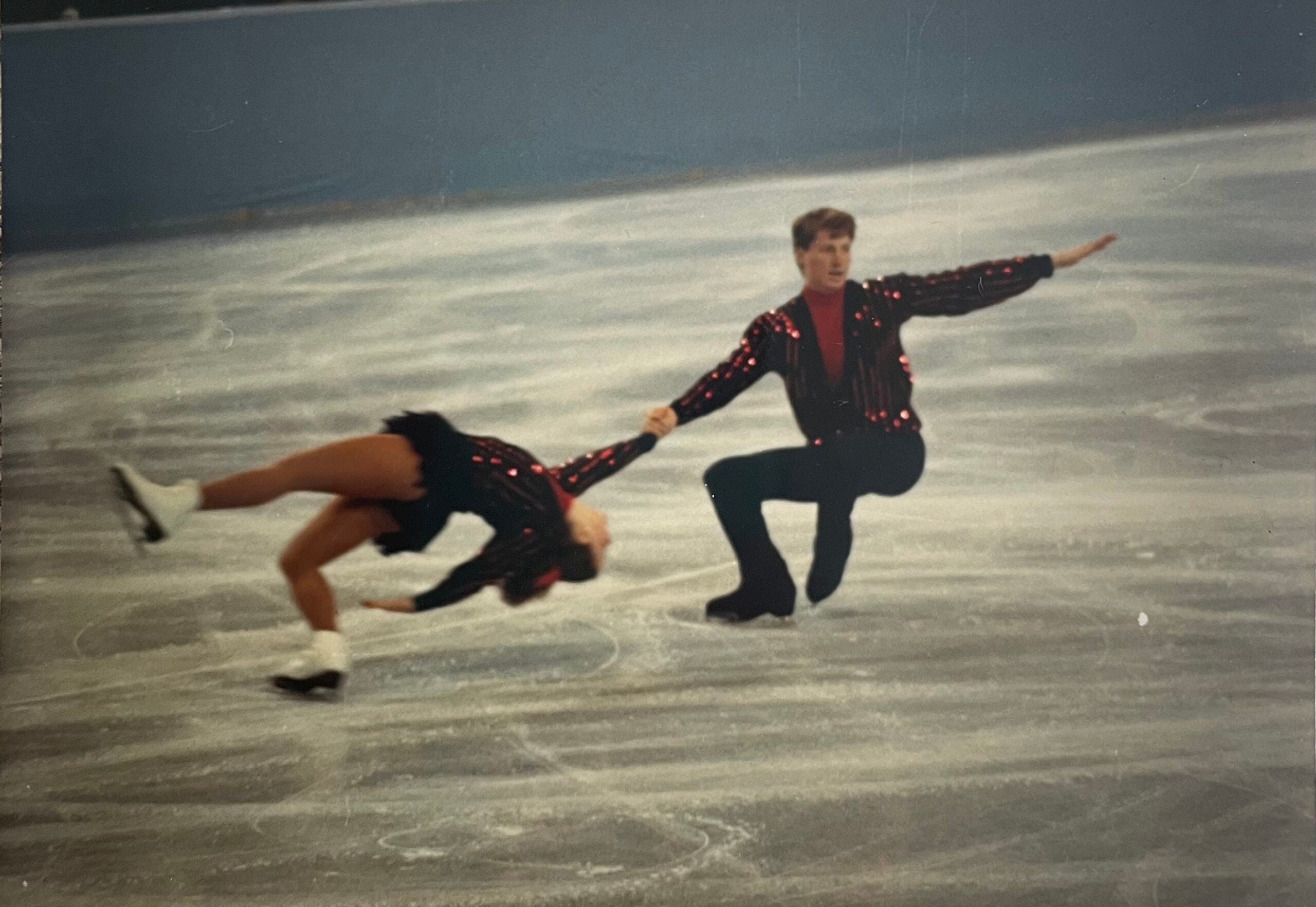 A pair of figure skaters perform a dramatic move on the ice, with the male skater supporting the female skater as she leans backward close to the ice.