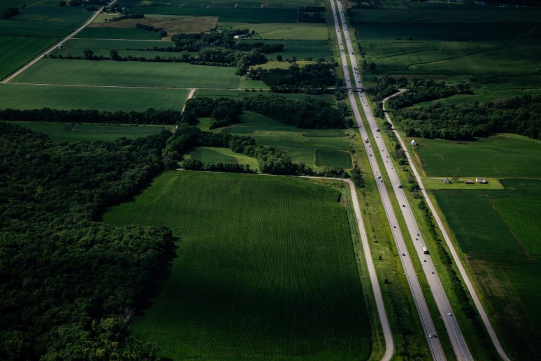 Aerial view of a highway running through green farmland and patches of forest, with vehicles traveling in both directions.