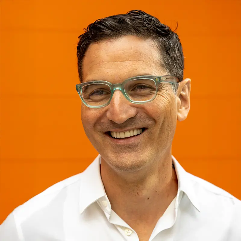 A man wearing clear-framed glasses and a white shirt smiles in front of an orange background.
