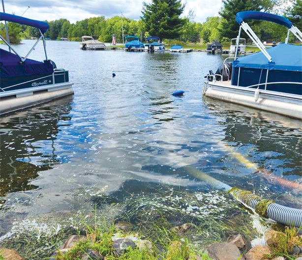 Two pontoon boats docked on a lake with hoses discharging foamy water near the shore; additional boats and trees are visible in the background.