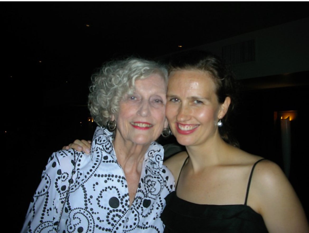 Two women are posing together, smiling at the camera. One woman has curly gray hair and is wearing a patterned blouse, while the other has long brown hair and is wearing a black dress.