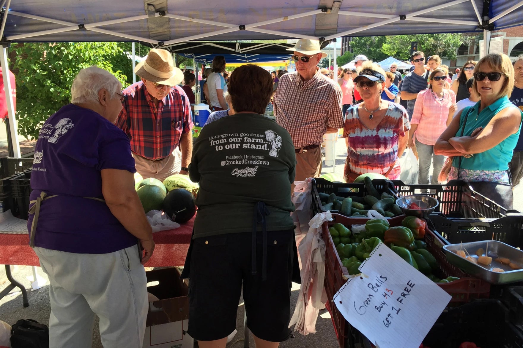 People shop for fresh produce at an outdoor farmers market stand featuring watermelons, peppers, and a Green Bells Buy 1 Get 1 FREE sign.