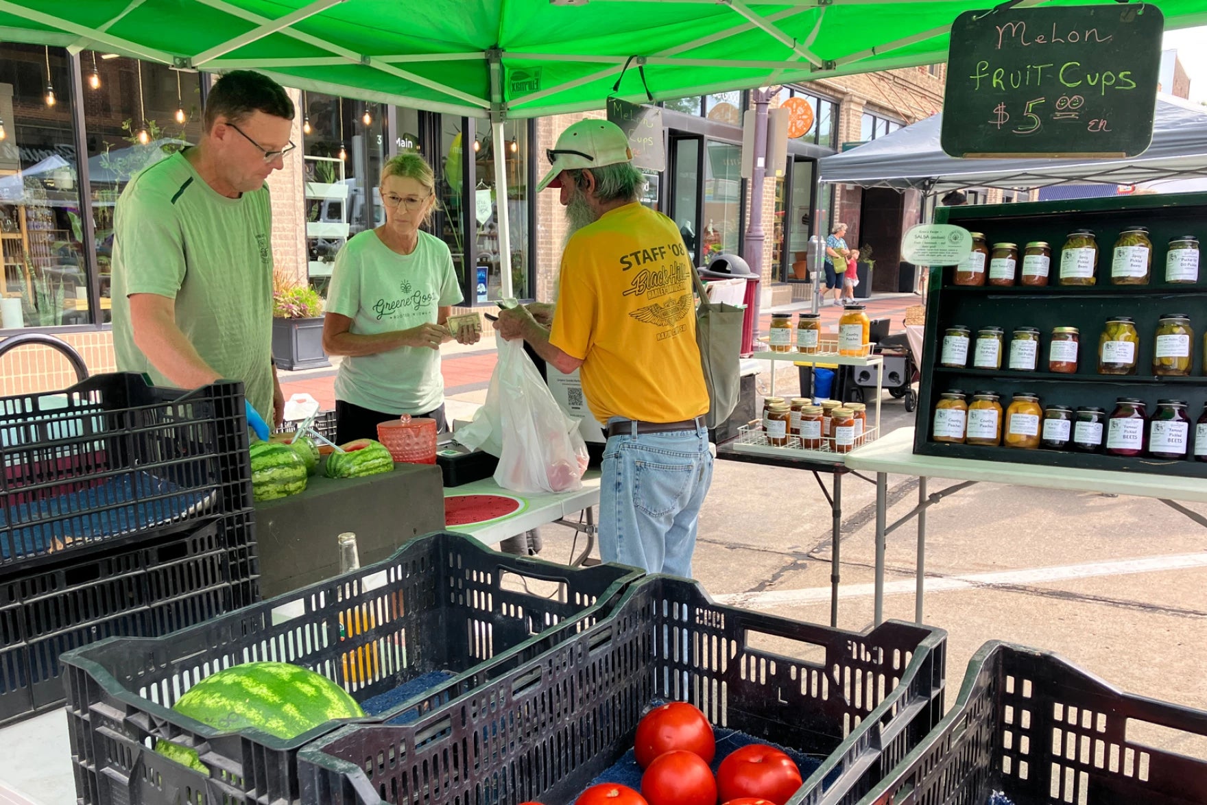 Three people interact at a farmers market produce stand with tomatoes, watermelons, jams, and a sign for $5 melon fruit cups.
