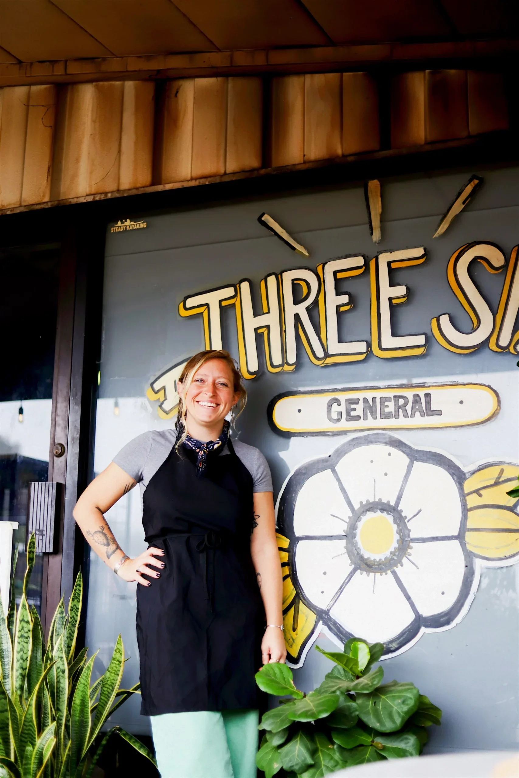 A woman wearing a black apron stands and smiles in front of a store window painted with the words THREE SIS and GENERAL, next to green plants.