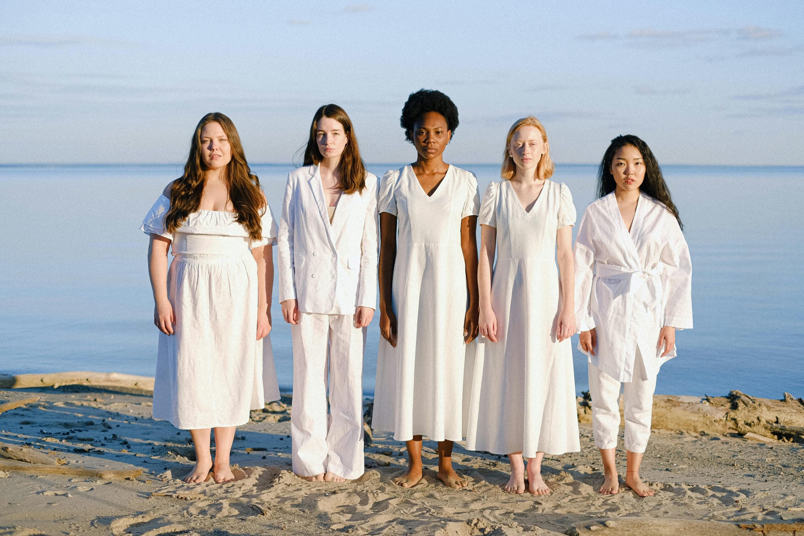 Five women stand barefoot on a sandy beach, facing the camera, dressed in different white outfits, with calm water and blue sky in the background.