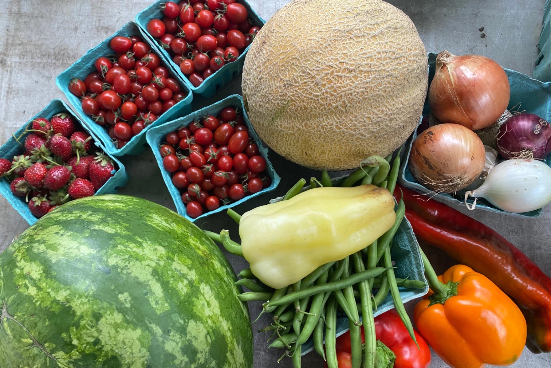 An assortment of fresh produce including cherry tomatoes, strawberries, cantaloupe, onions, watermelon, green beans, bell peppers, and long red peppers on a table.