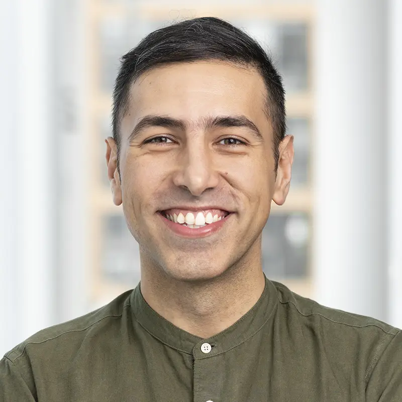 A man with short dark hair, wearing a green button-up shirt, smiles while standing indoors with blurred windows in the background.