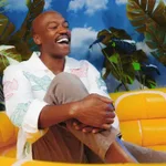 A man sits on a yellow couch, smiling and laughing, with tropical plants and a blue sky background behind him.