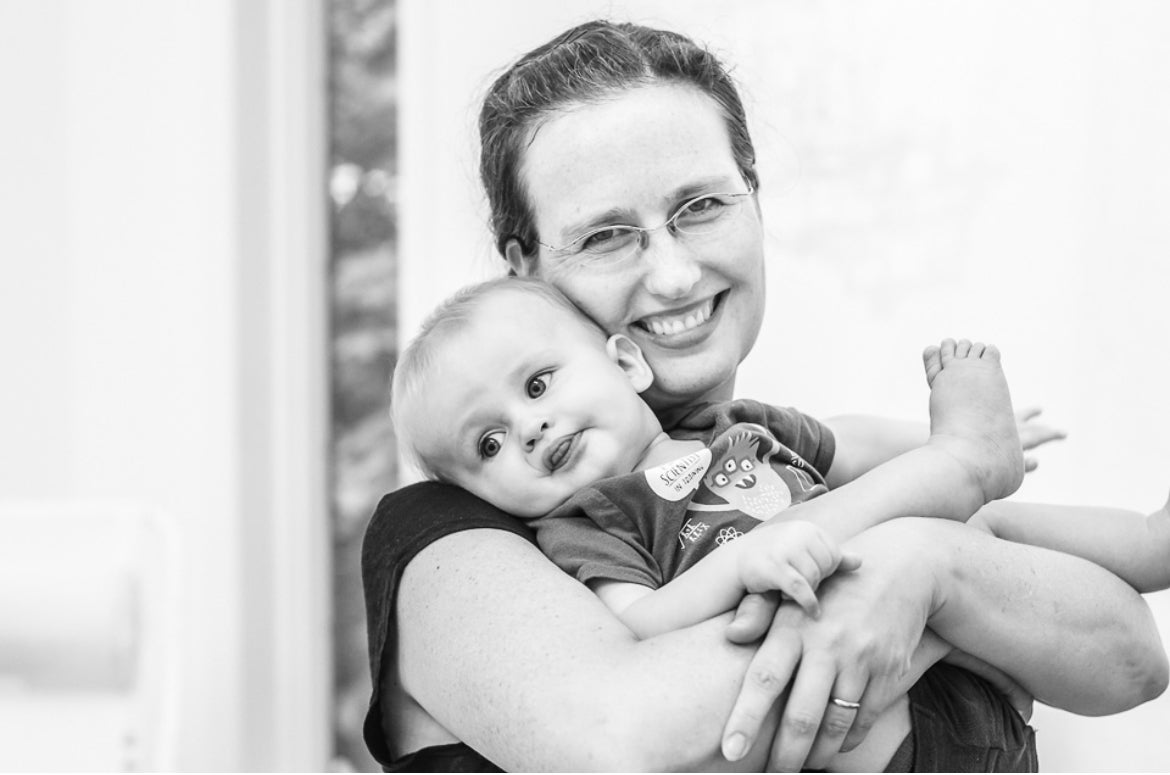 A woman wearing glasses smiles while holding a baby who is resting in her arms. Both appear happy and are looking toward the camera in a bright, indoor setting.