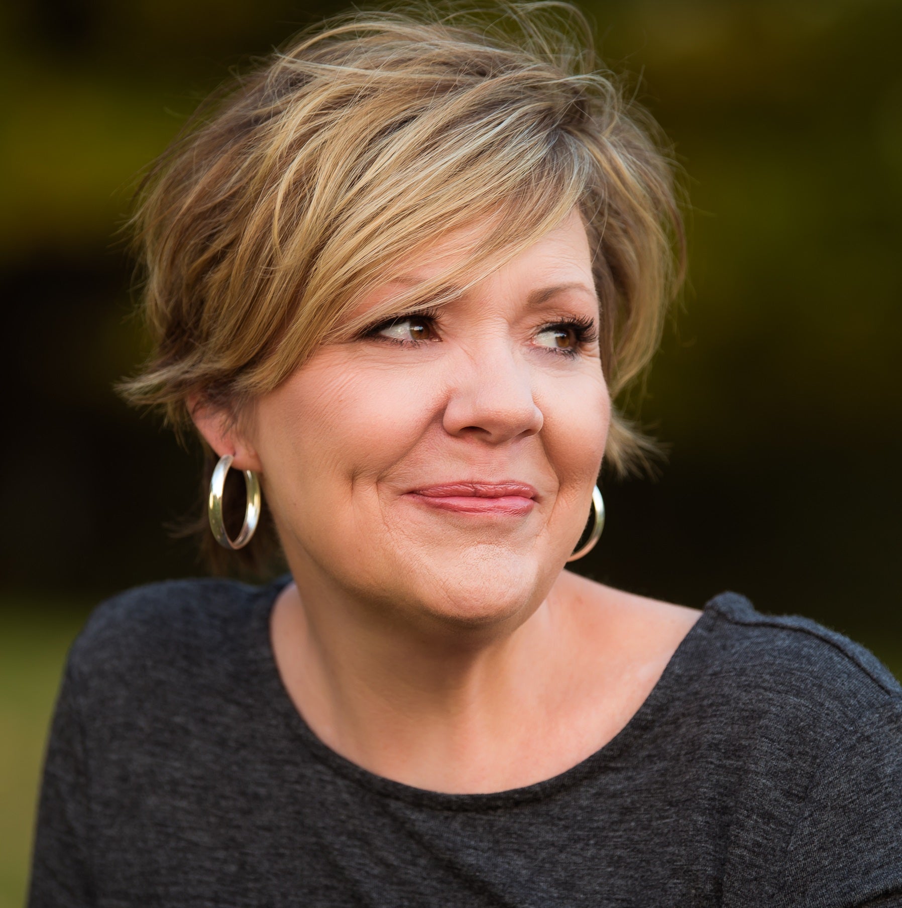 A woman with short blonde hair and hoop earrings, wearing a dark shirt, looks to the side and smiles softly outdoors.