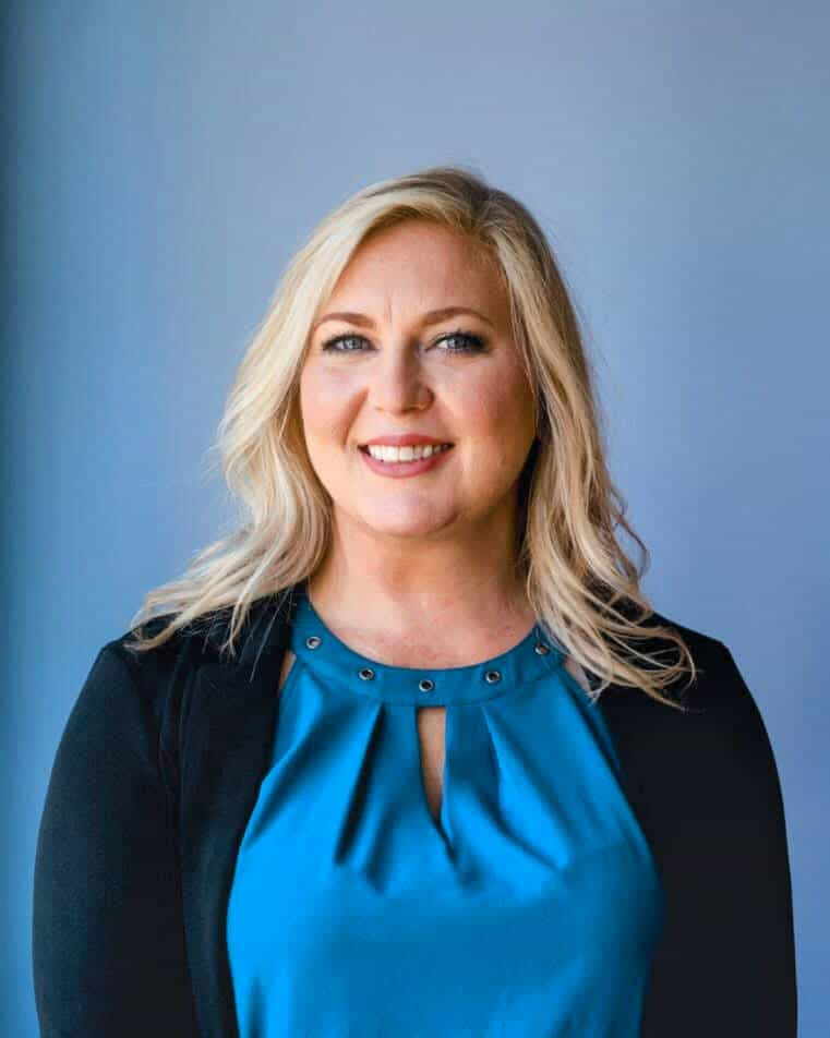 A woman with long blonde hair wearing a blue blouse and black cardigan stands in front of a plain blue background, smiling at the camera.