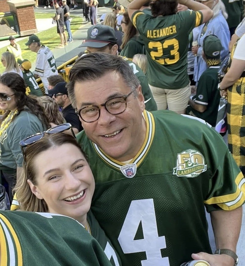 Two people wearing Green Bay Packers jerseys smile for a selfie at an outdoor event among a crowd of other Packers fans.