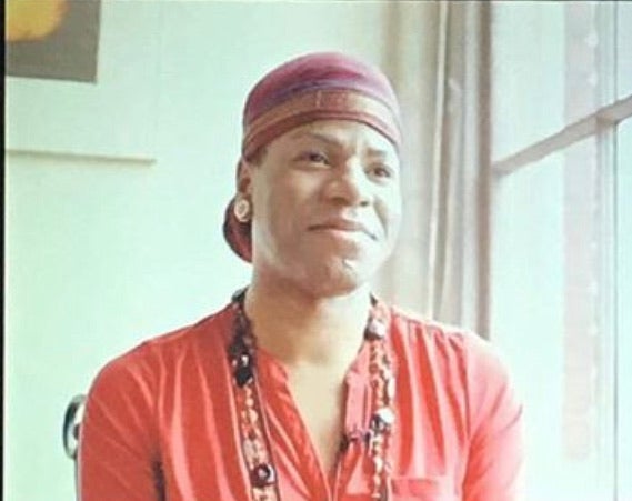 Person wearing a red headscarf and red shirt with a beaded necklace sits indoors, facing slightly to the right with a neutral expression.