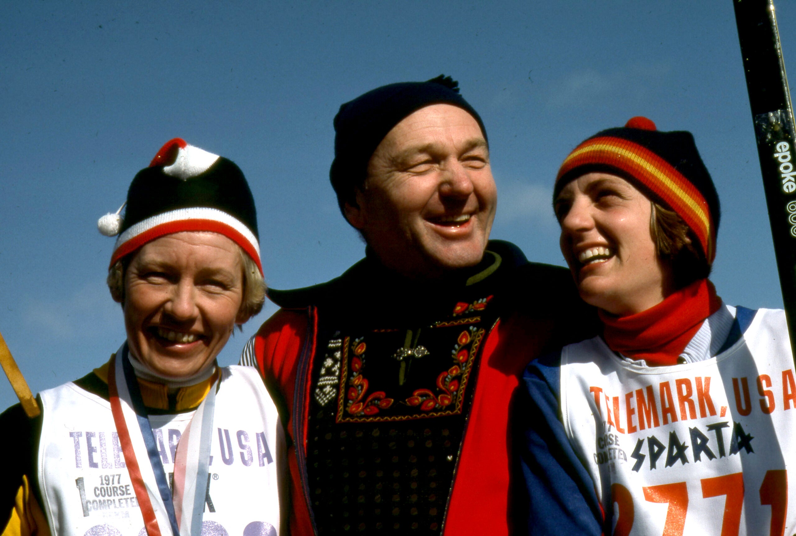 Three people wearing winter sports clothing and bibs smile outdoors, with one person in traditional clothing standing between two skiers.