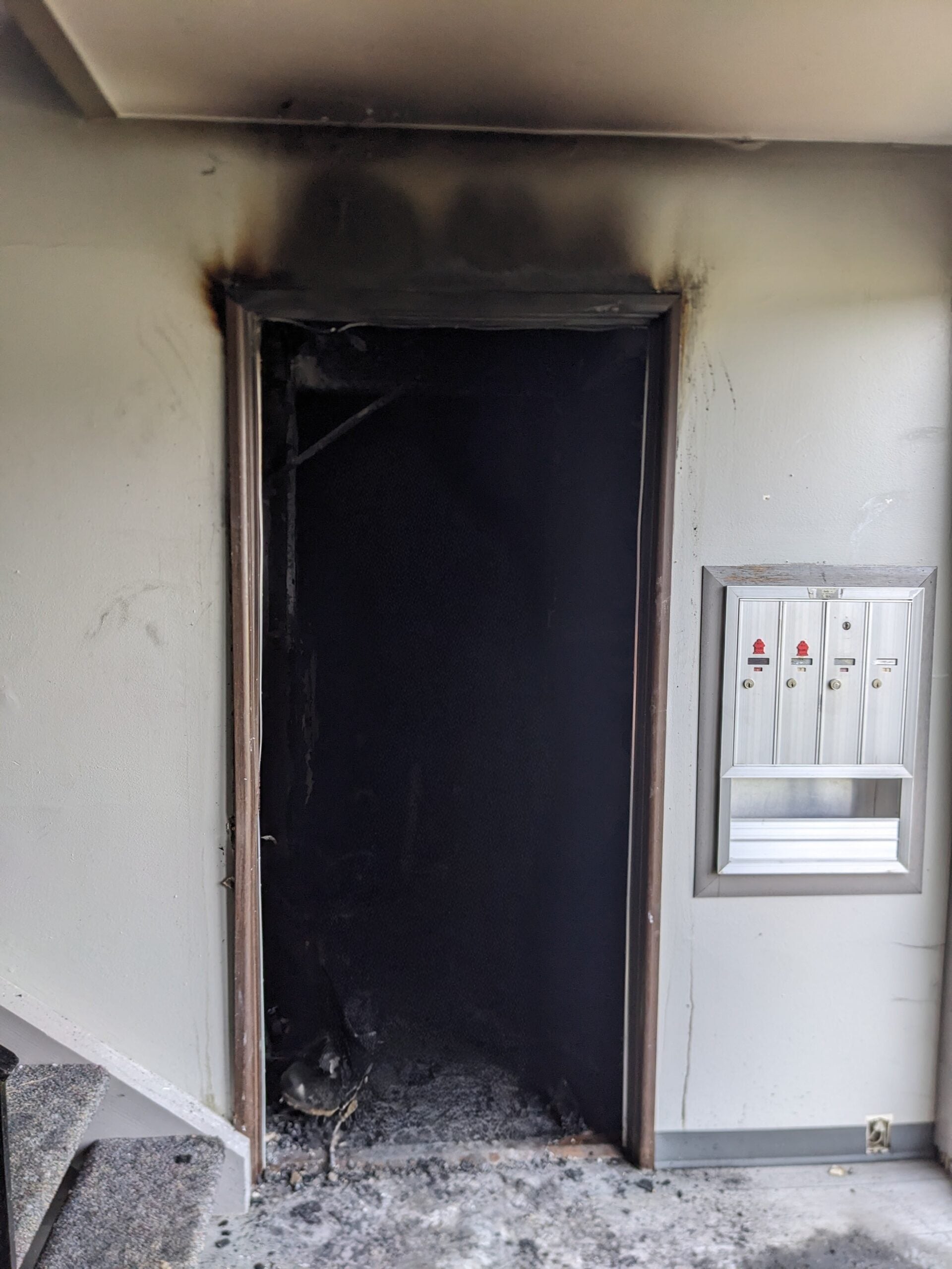 A doorway with severe fire damage, including blackened walls and ceiling above the door; nearby wall-mounted controls remain mostly unaffected.