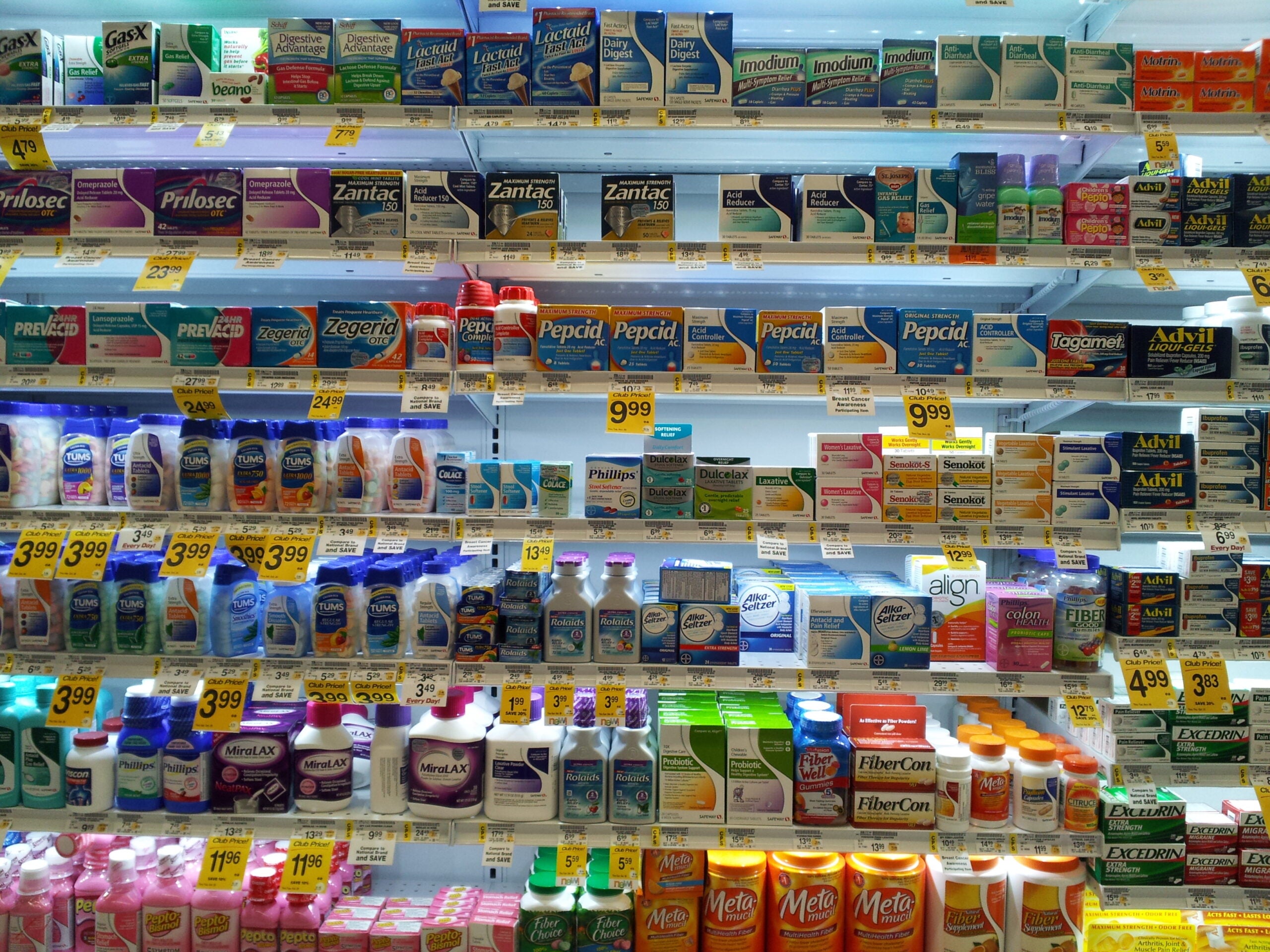 Shelves stocked with various over-the-counter medications and supplements, including antacids, pain relievers, digestive aids, and laxatives, with price tags visible.