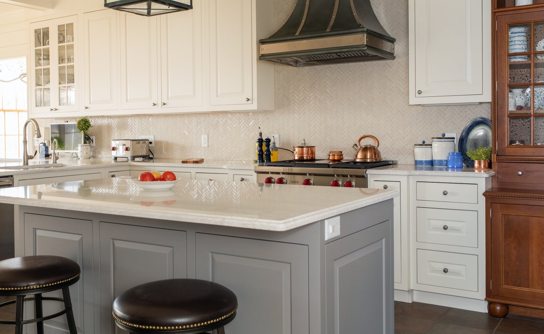 Modern kitchen with white cabinets, a gray island, black range hood, and stainless steel appliances. Three red apples sit on the island countertop with two stools in front.