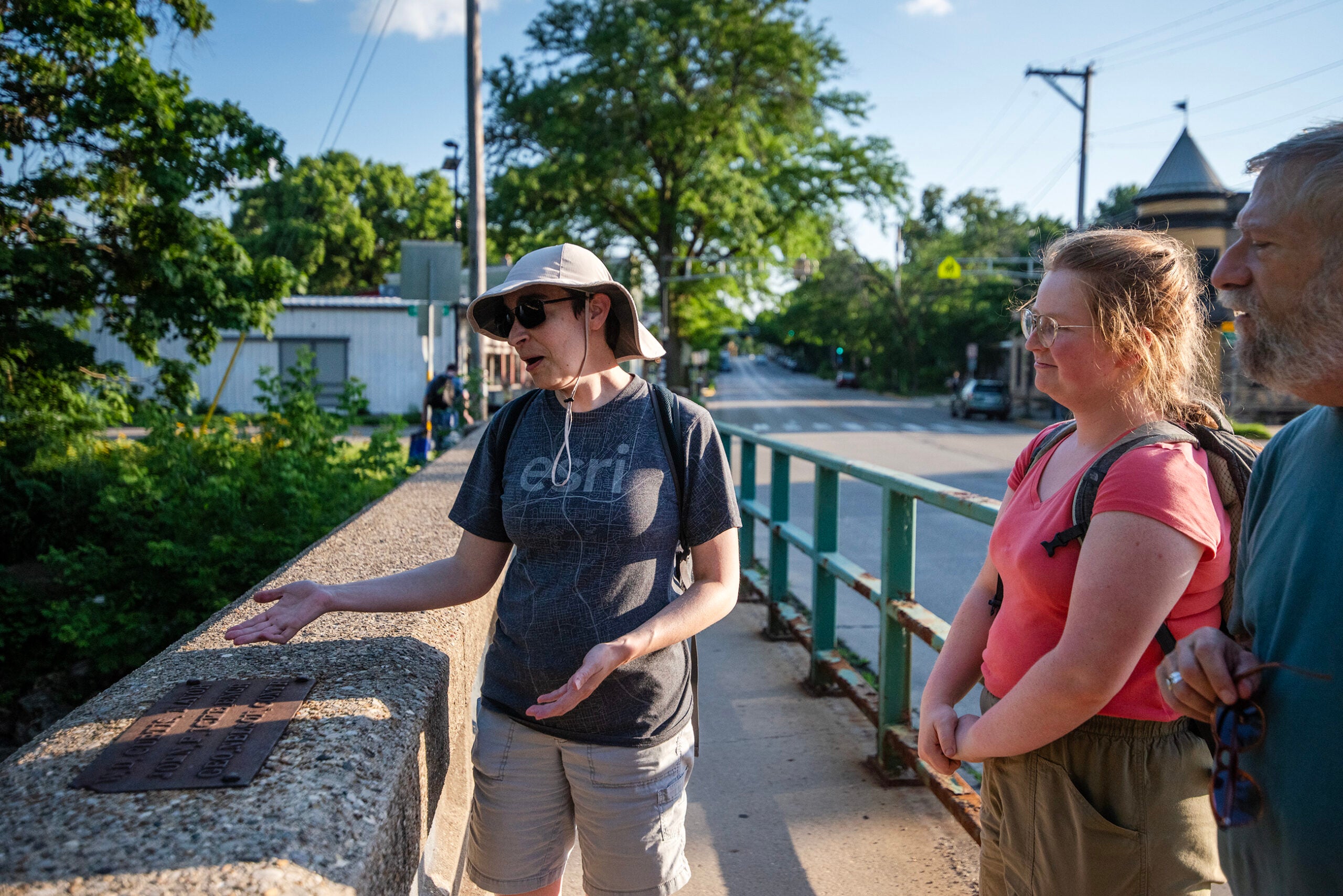 A person gestures toward a plaque on a bridge while two others watch, with trees and a road in the background on a sunny day.