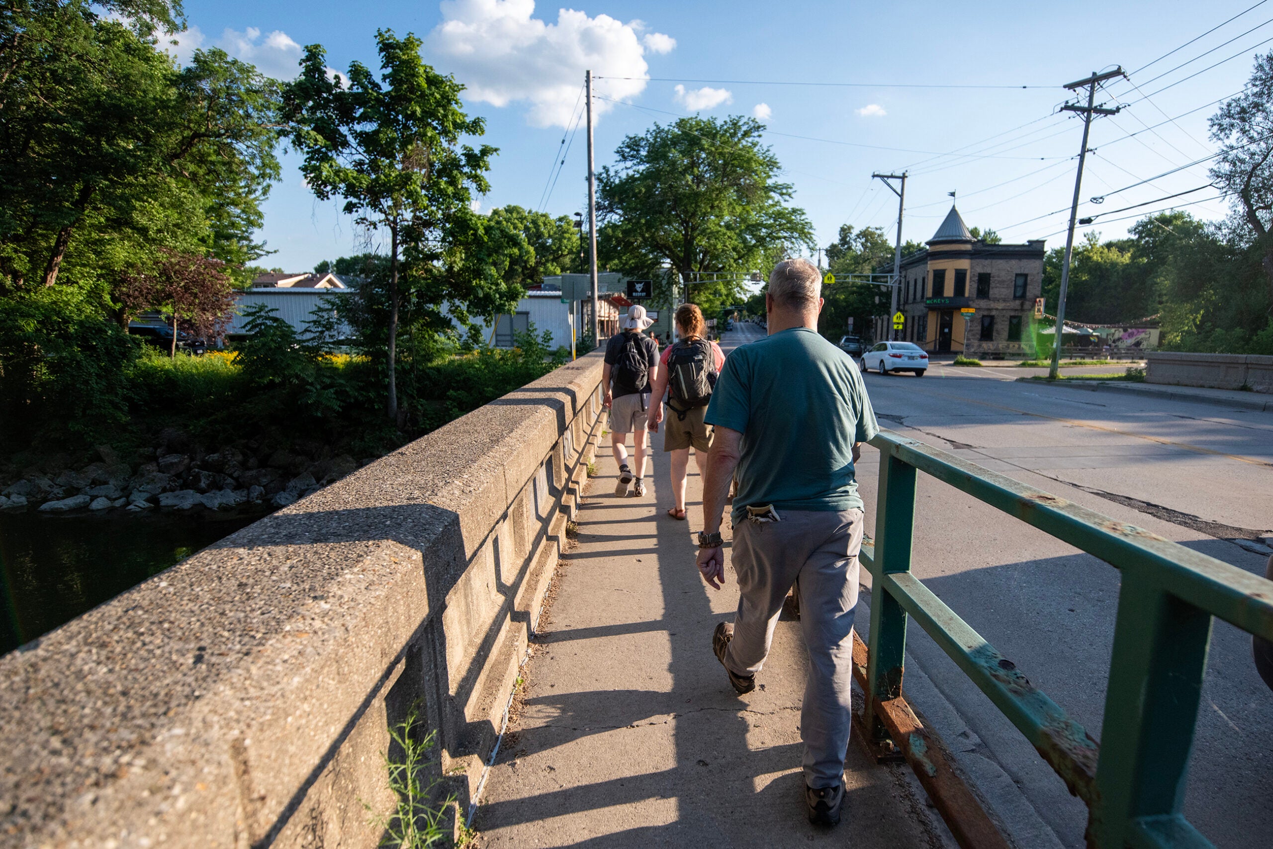A group of people walk together on a sidewalk alongside a bridge on a sunny day, with trees and buildings in the background.