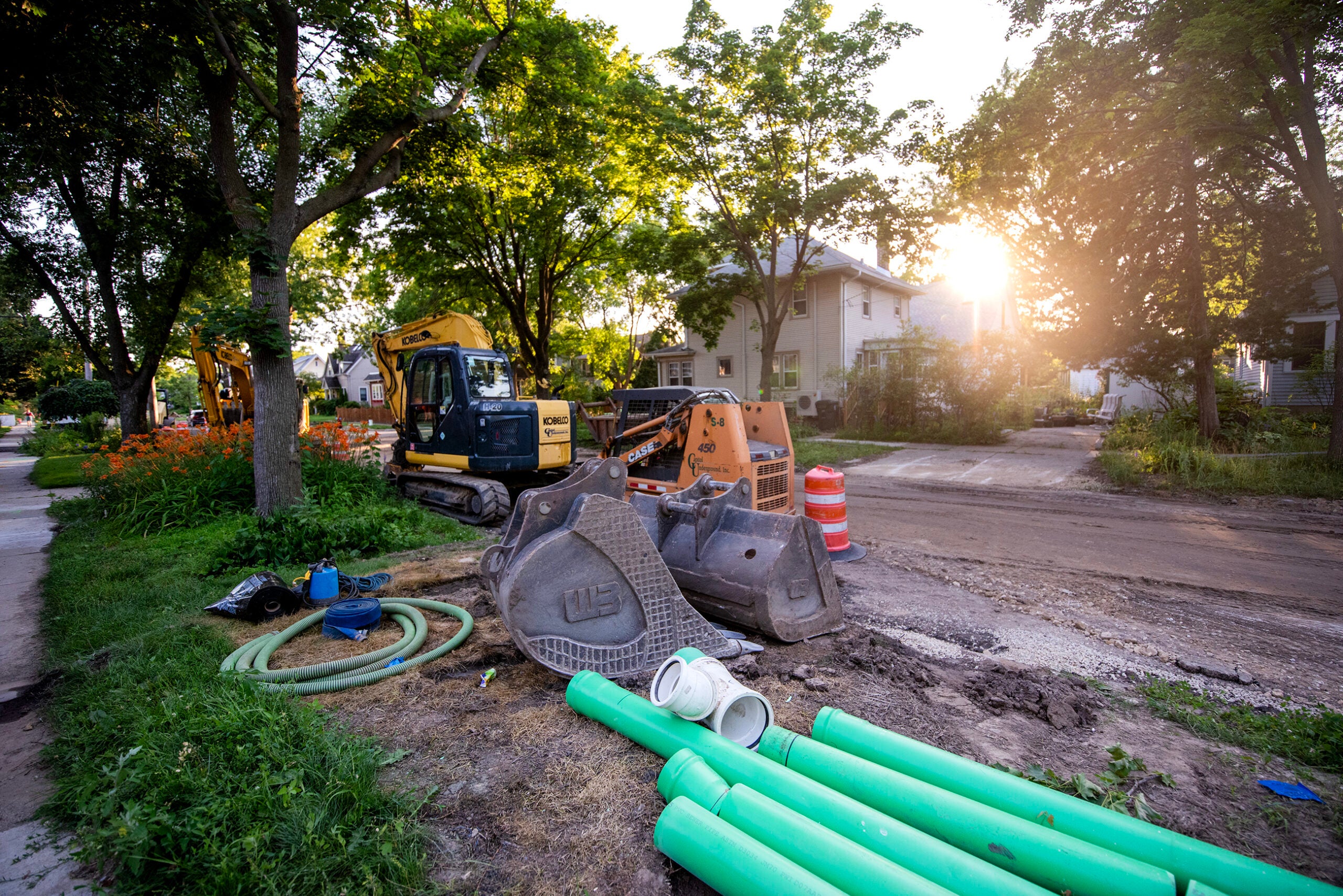 Construction site on a residential street with excavators, green pipes, traffic barrels, hoses, and construction equipment; sunlight shines through trees in the background.