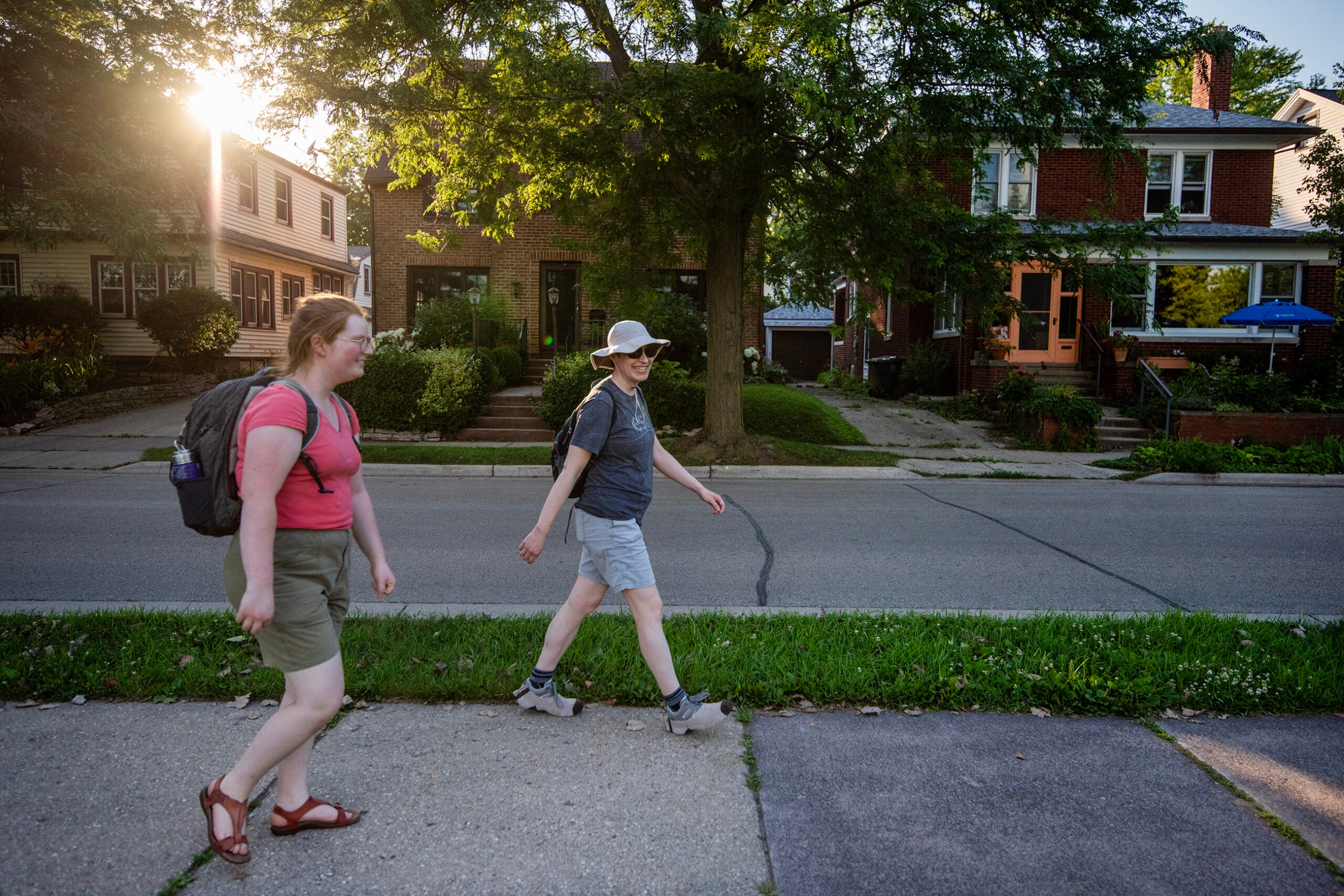 Two people with backpacks walk on a sidewalk in a residential neighborhood with houses and trees, during daylight.
