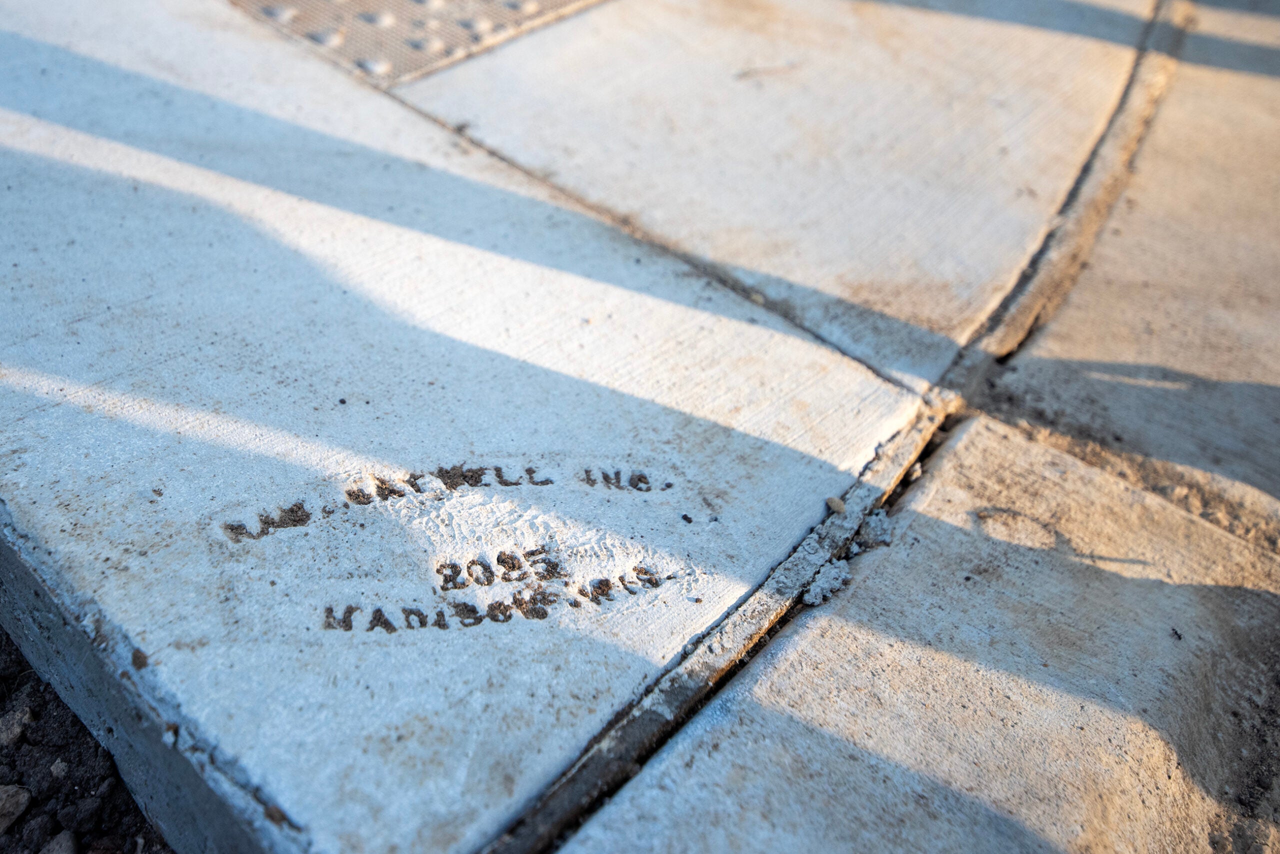 A close-up of a concrete sidewalk corner with an imprint reading RMS MAXWELL INC. 2023 MAN366-PR5 partially visible on the surface.
