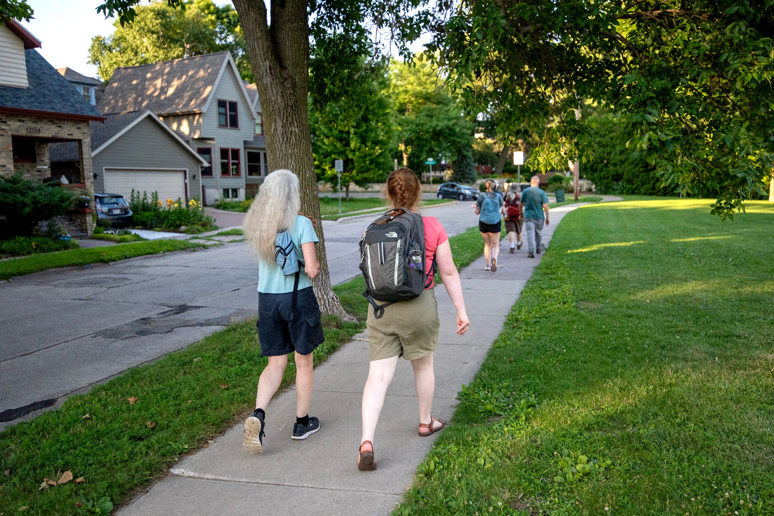 A group of people walk along a suburban sidewalk next to houses and a grassy area on a sunny day.