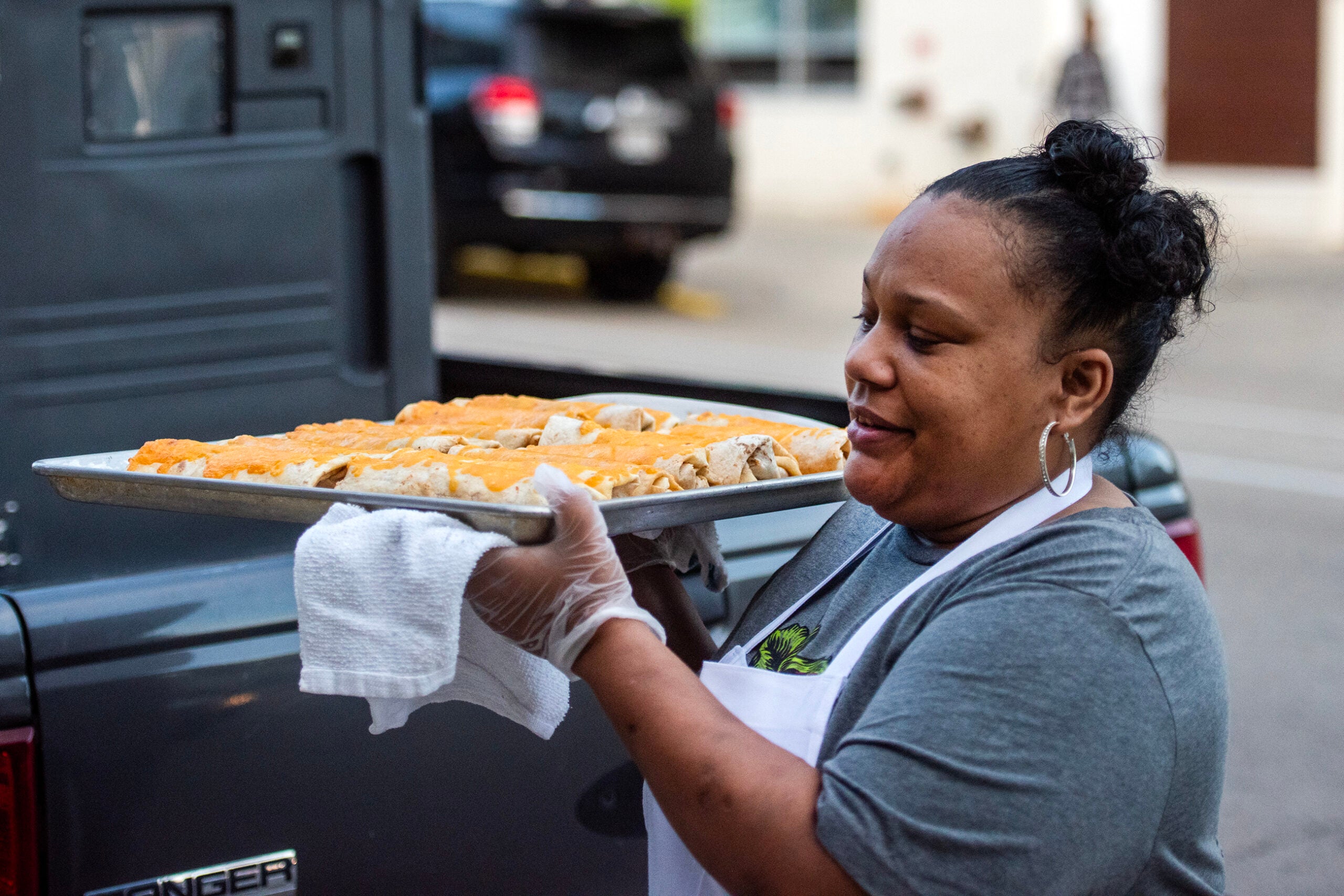 A woman wearing an apron and gloves holds a tray of food in an outdoor setting near a parked vehicle.