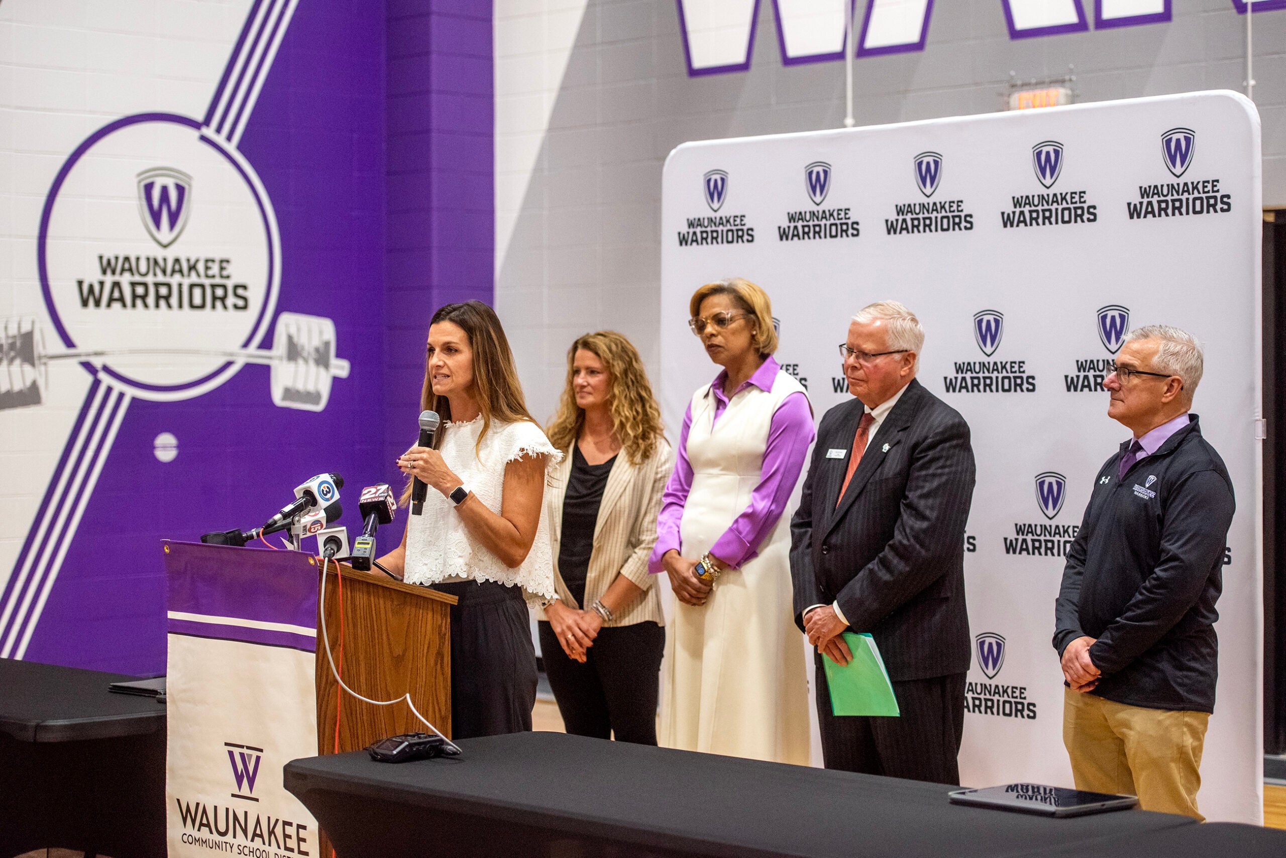 A group of five adults stand near a podium and microphones in a gymnasium with Waunakee Warriors branding visible on the wall and banner.