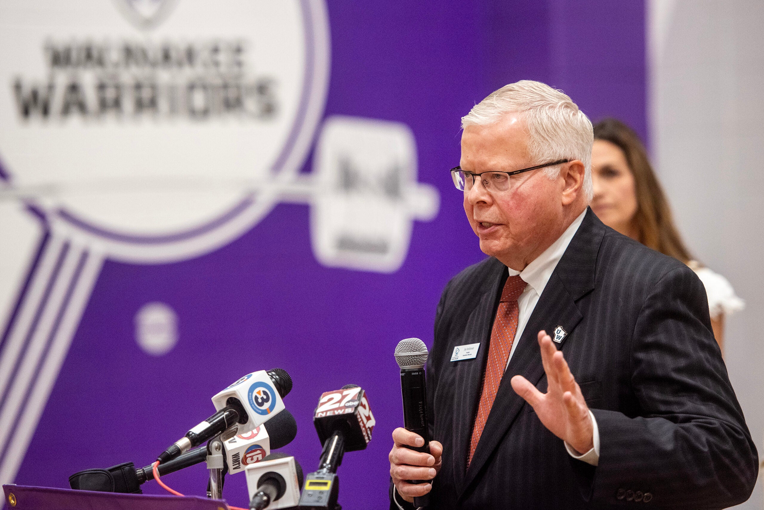 An older man in a suit speaks at a podium with multiple microphones, in front of a purple backdrop that says Milwaukee Warriors.
