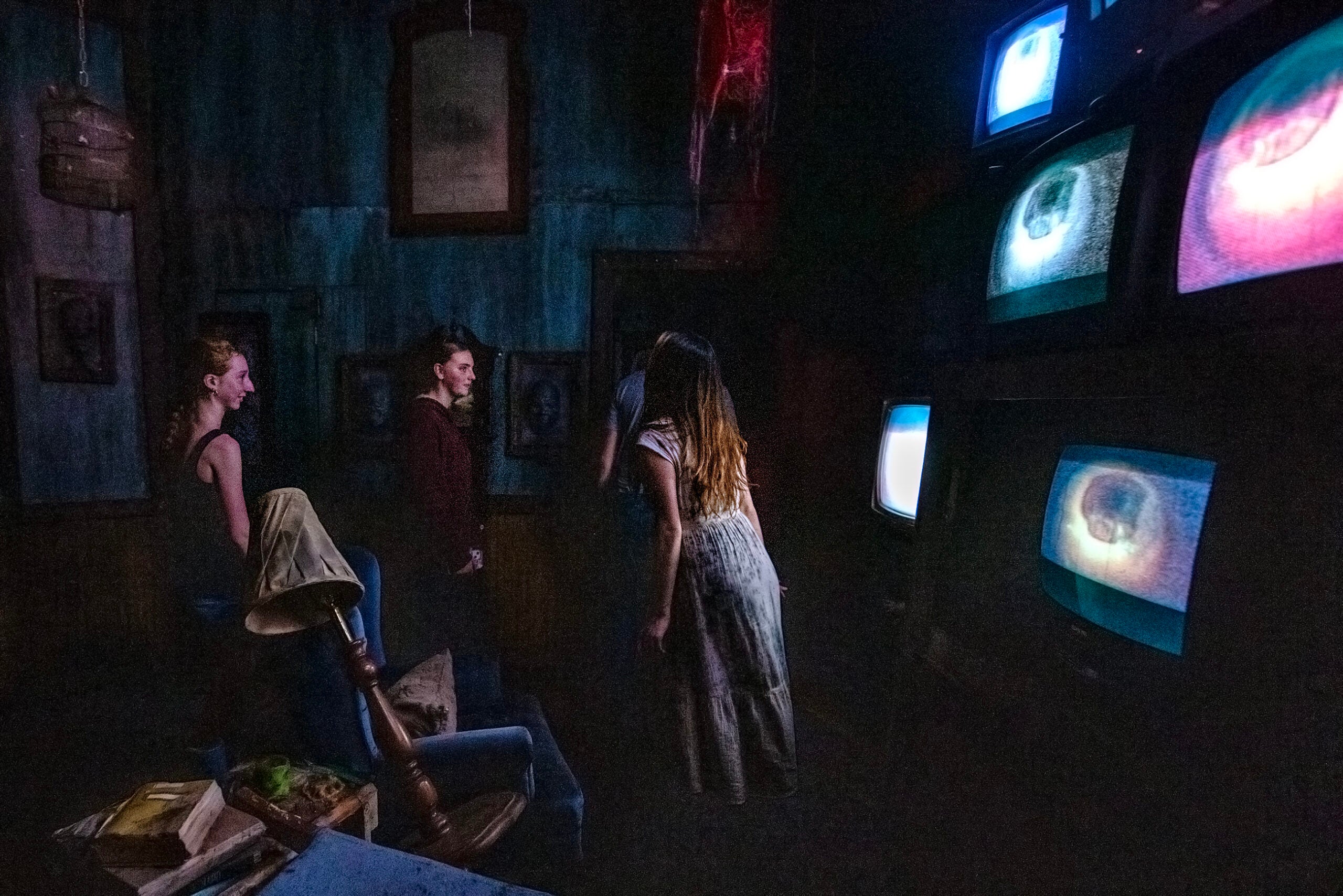 Four people stand in a dimly lit room, watching large screens displaying close-up images of eyes; old furniture and portraits are visible around them.