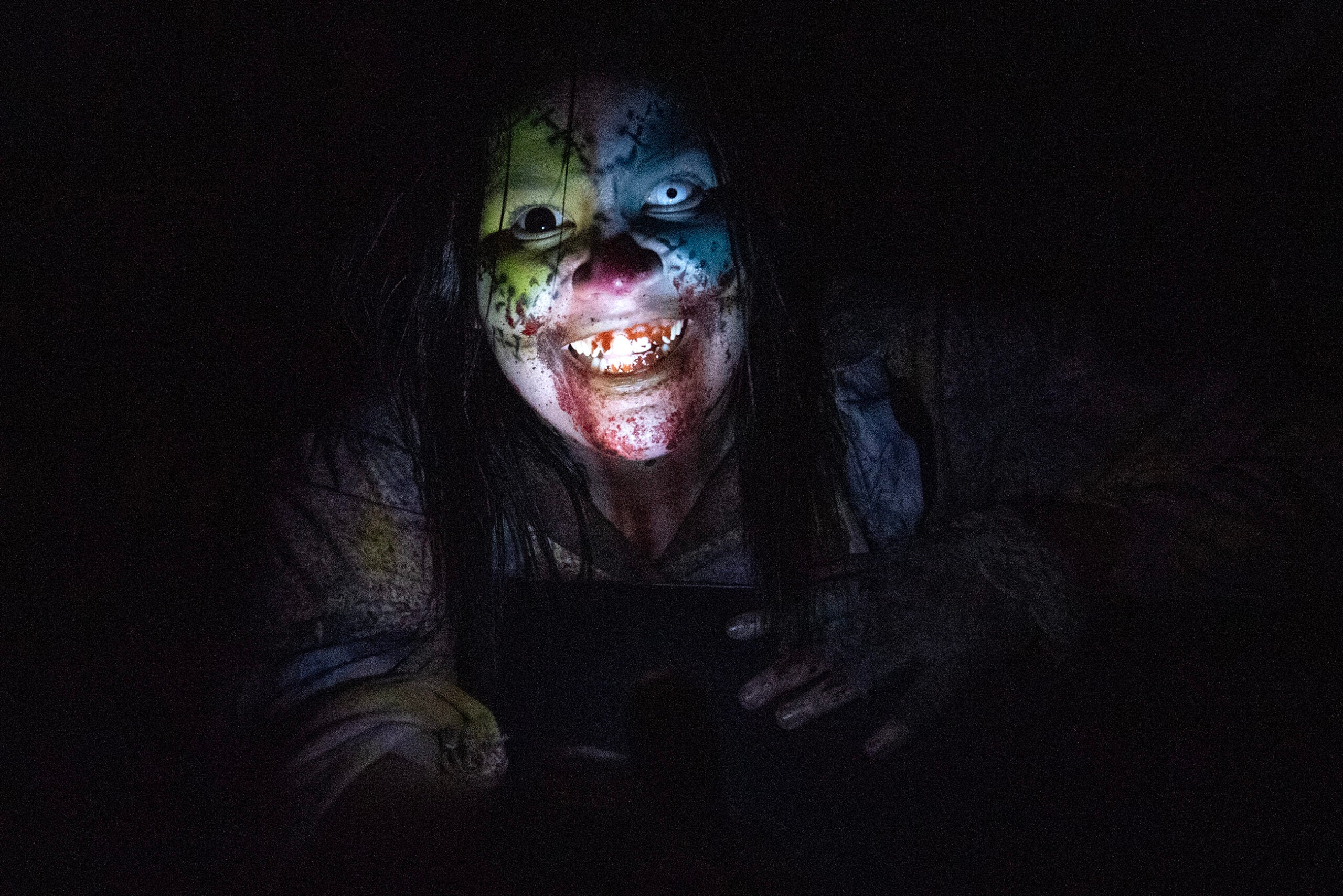 Person in dark setting with long hair and painted clown face, showing sharp teeth and illuminated from below, wearing tattered clothing.