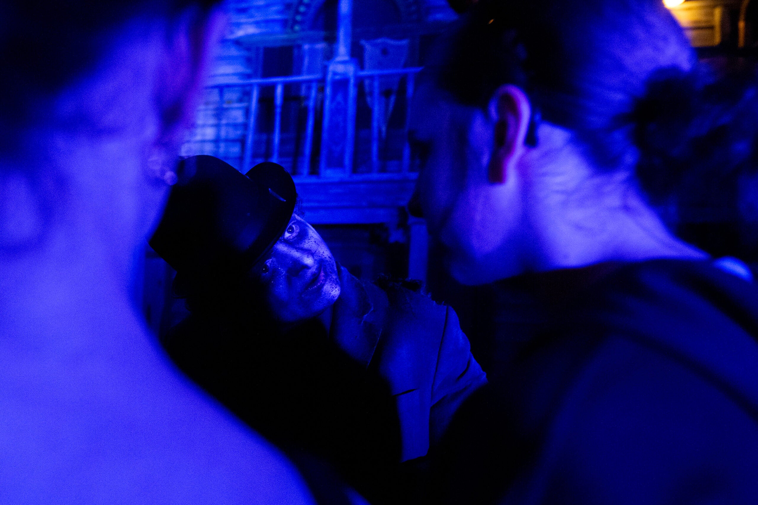 A person in a hat illuminated by blue light looks intently at two other people in the foreground, with a balcony and building in the background.