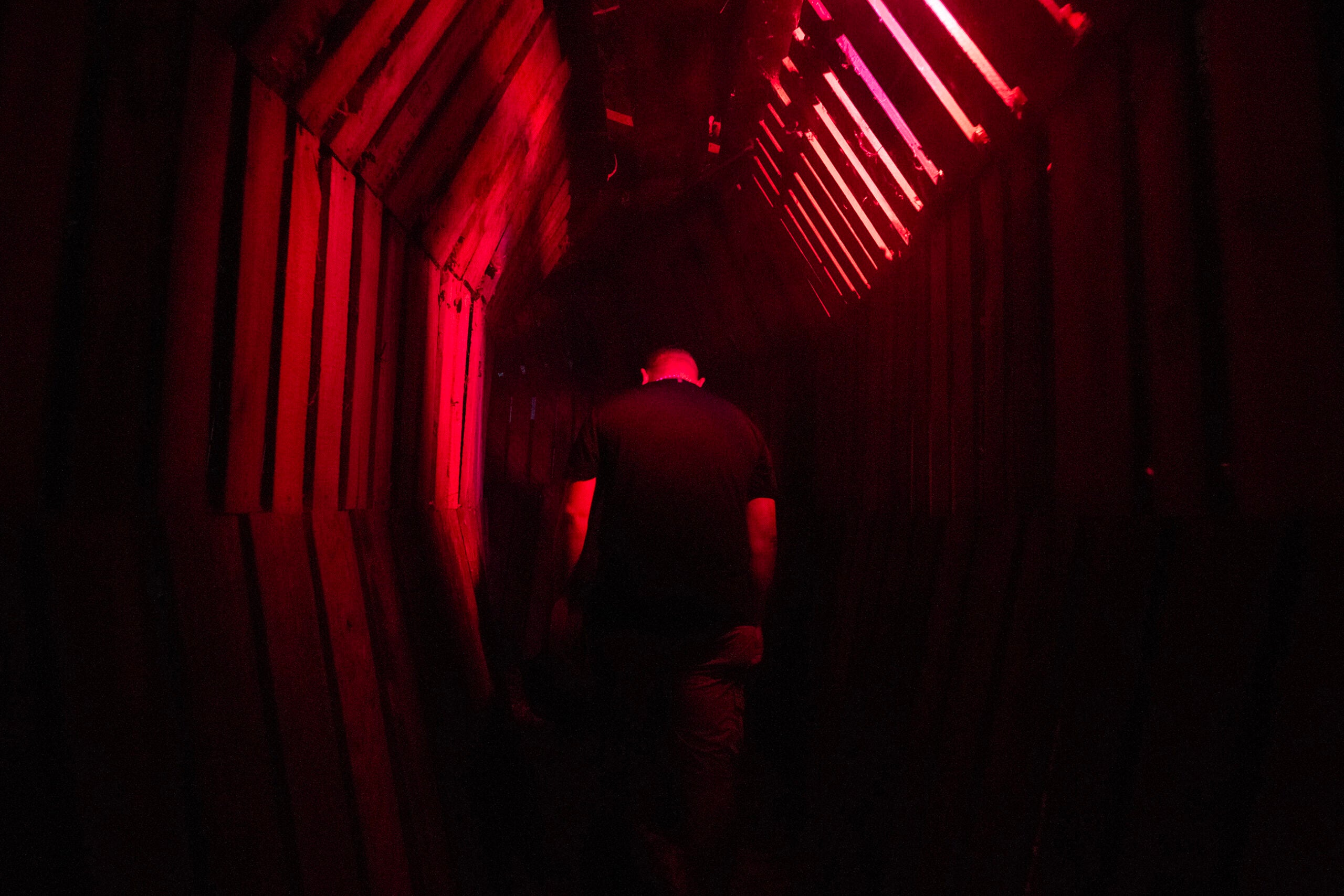 A person walks through a narrow, dimly lit corridor with red lighting and wooden walls.