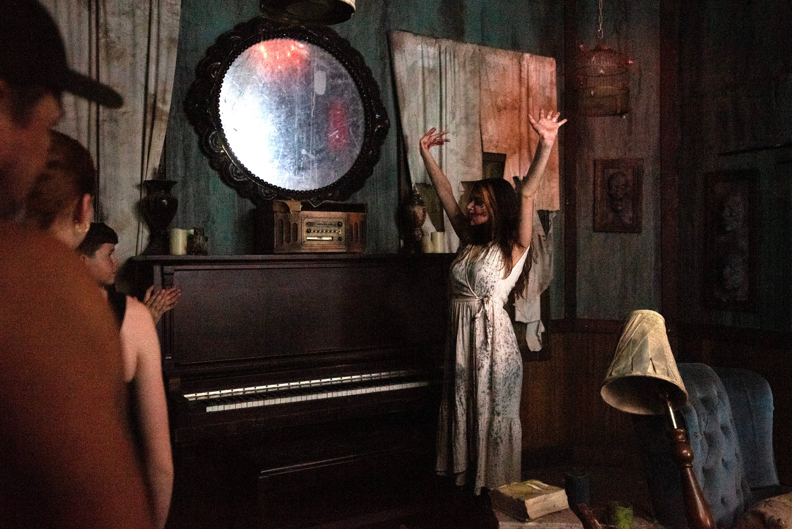 A woman in a white dress stands with her arms raised in front of an upright piano, while several people watch her in a dimly lit, old-fashioned room.