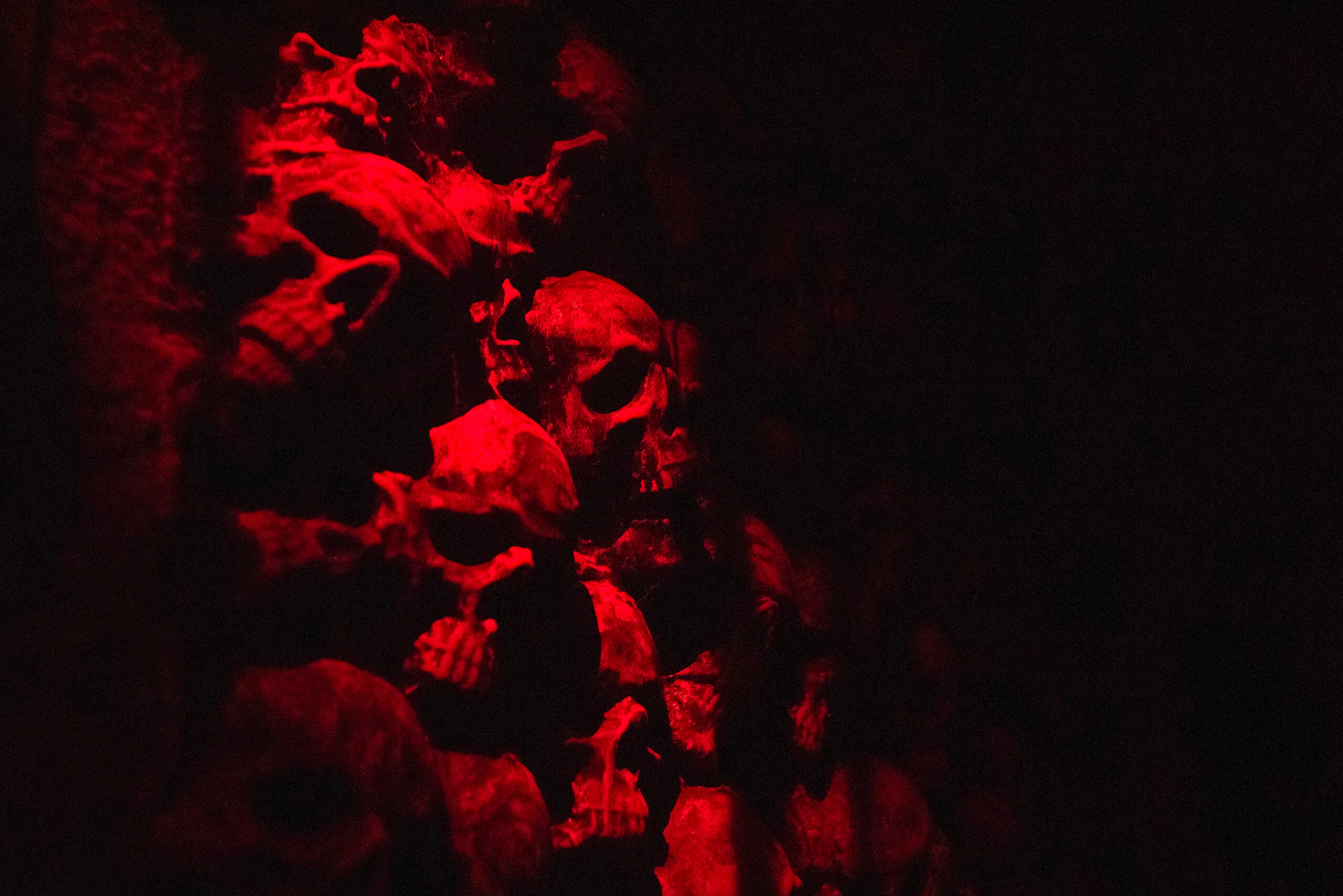 A cluster of human skulls stacked closely together, illuminated by dim red lighting against a dark background.