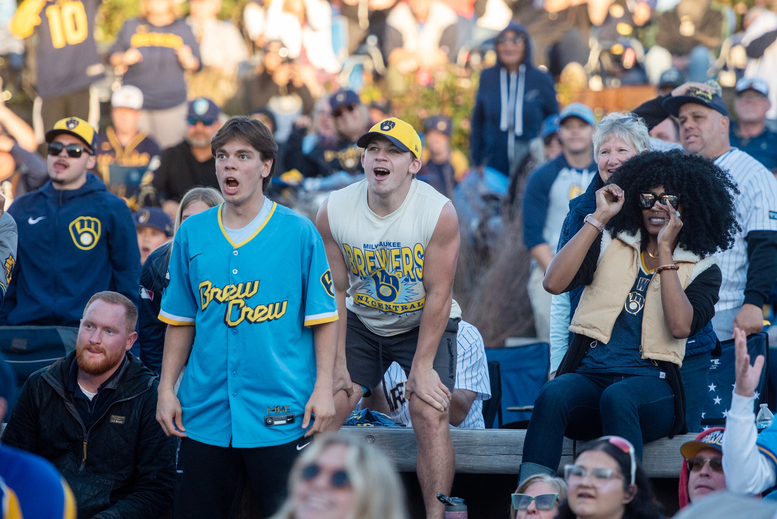 A crowd of Milwaukee Brewers fans watch a game outdoors, with some fans standing and reacting with excitement or surprise.