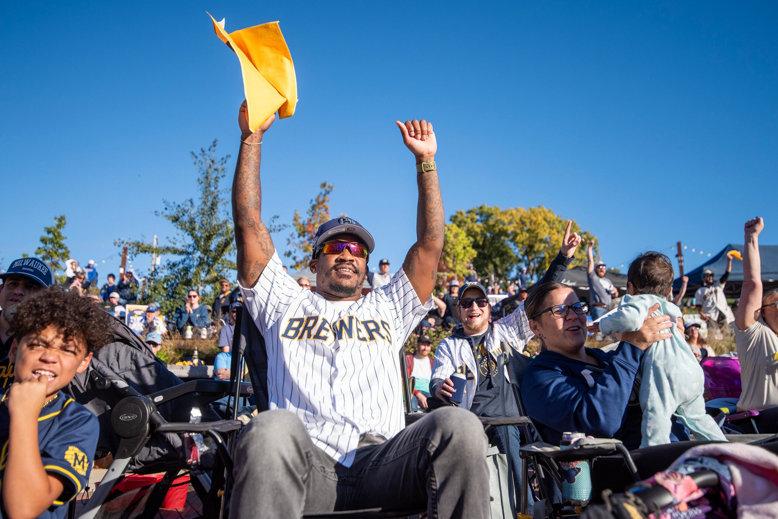 A crowd of people outdoors, many wearing Milwaukee Brewers gear, cheer and raise their hands while watching an event on a sunny day.