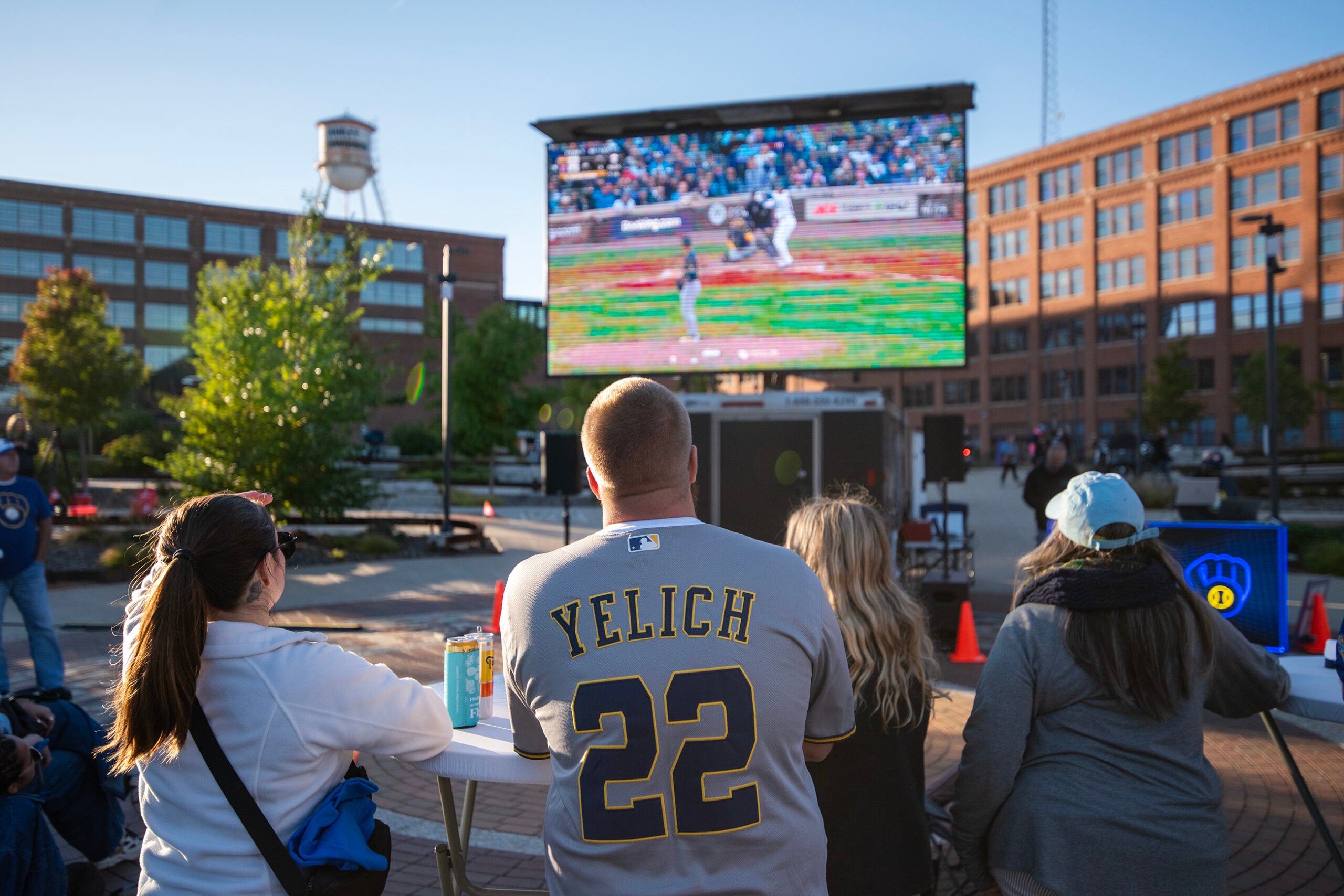A group of people sit at a table outdoors watching a baseball game on a large screen, with buildings and a water tower in the background.